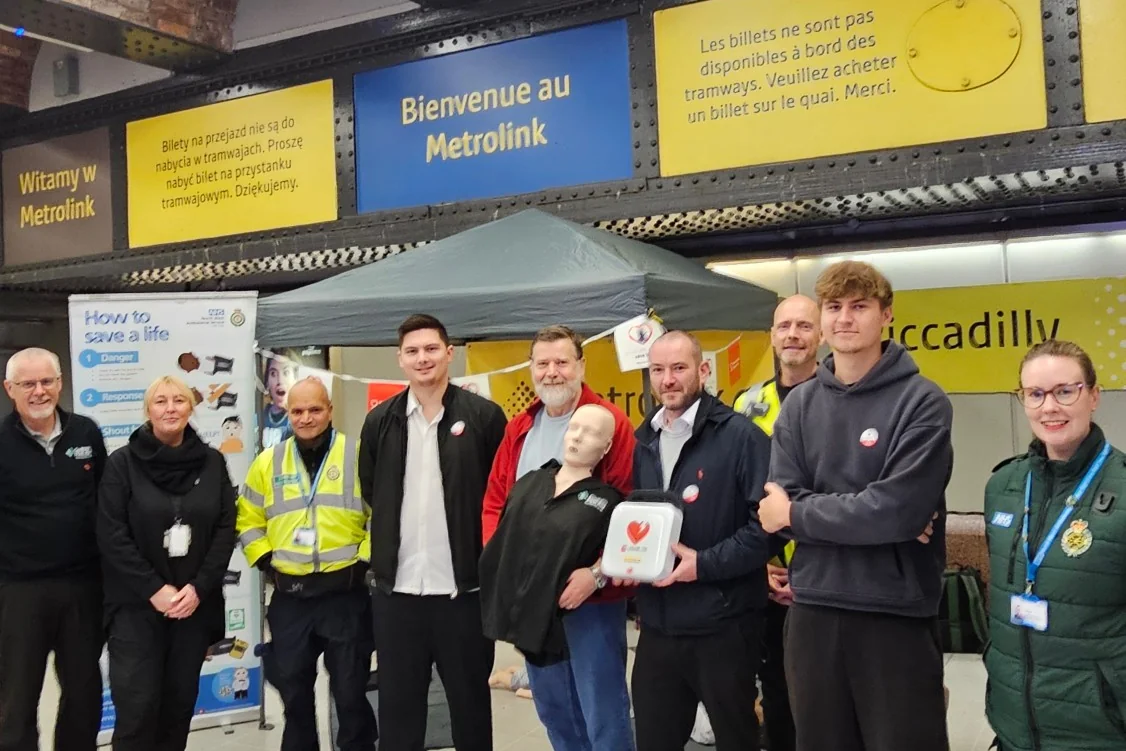 The Oxylitre team with ambulance service and Metrolink staff at a Restart a Heart CPR training stand in Piccadilly Metrolink station, holding a training manikin and a defibrillator